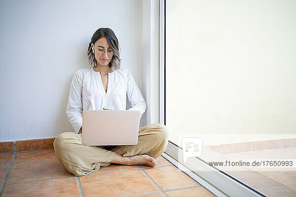 Freelancer sitting cross-legged using laptop by window at home