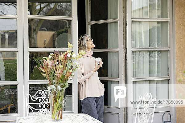Thoughtful senior woman holding coffee cup standing at doorway