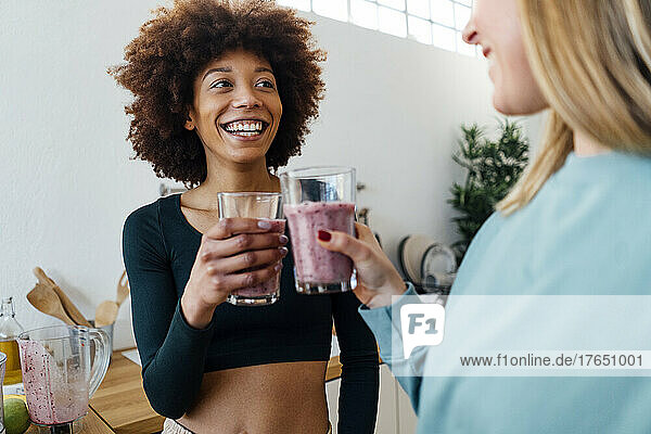 Cheerful young woman toasting glass of smoothie with friend in kitchen