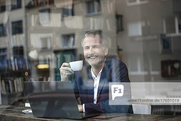 Happy freelancer with coffee cup and laptop seen through glass window at cafe
