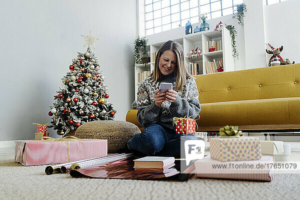 Glückliche Frau  die ihr Mobiltelefon benutzt und zu Hause neben Geschenken sitzt