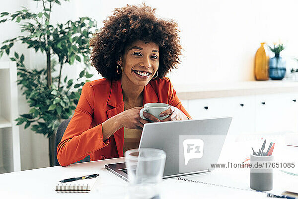 Smiling businesswoman holding coffee cup sitting with laptop at desk