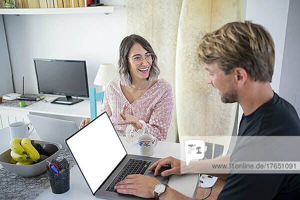 Happy young freelancer gesturing discussing with woman using laptop sitting at dining table