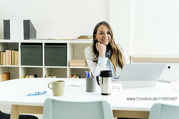 Smiling businesswoman with laptop sitting hand on chin at desk in office