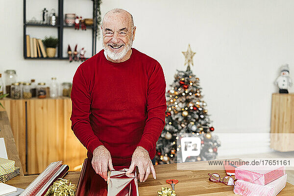 Fröhlicher älterer Mann verpackt Weihnachtsgeschenke auf dem heimischen Tisch