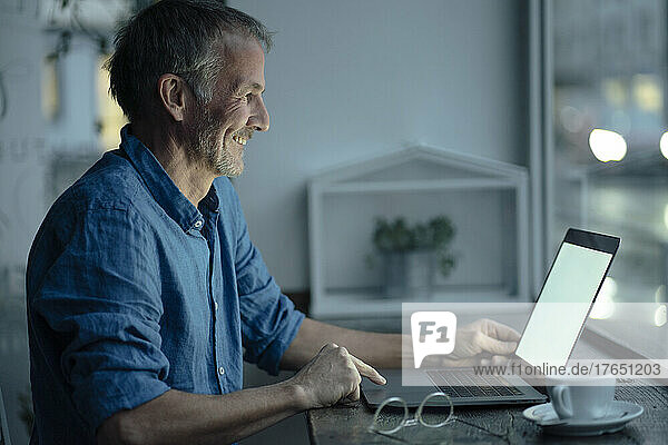 Smiling freelancer using laptop sitting at table by glass window in cafe