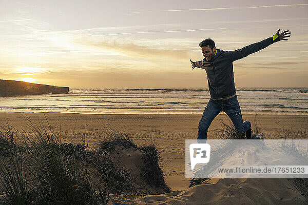 Carefree man jumping from sand dune with arms outstretched at beach