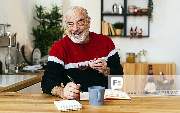Happy senior man holding eyeglasses sitting with diary at table at home