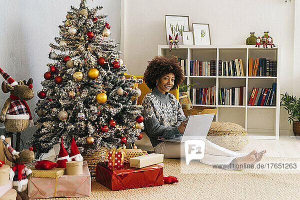 Happy young woman with laptop sitting by Christmas tree at home
