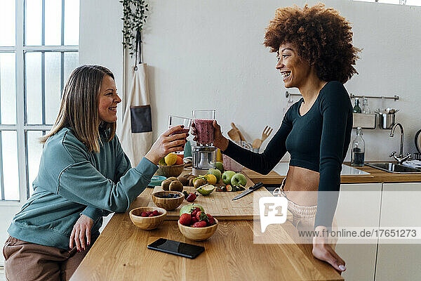 Happy young woman toasting glass of smoothie with friend sitting at table in kitchen