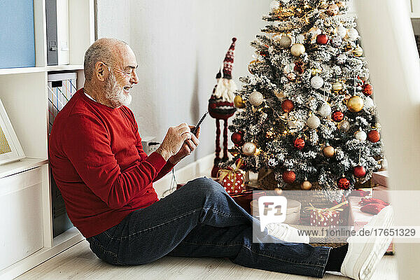 Happy senior man using smart phone sitting by Christmas tree at home