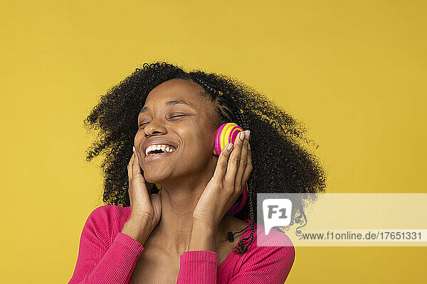Happy young woman with eyes closed enjoying music through wireless headphones against yellow back ground