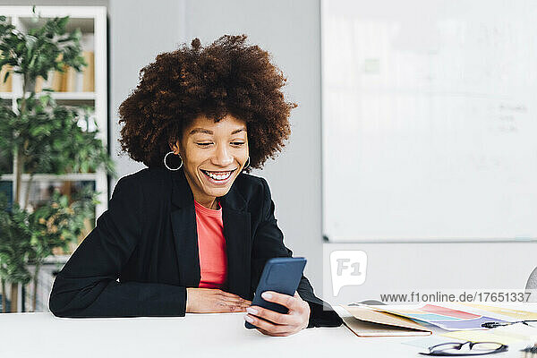 Happy businesswoman sitting at desk and using mobile phone in office