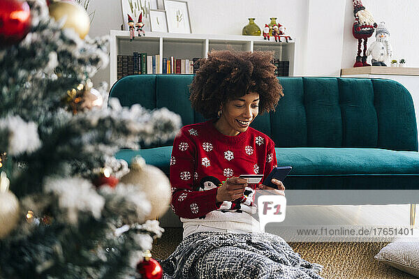 Happy woman with mobile phone and credit card sitting by sofa at home