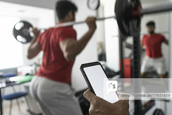 Hand of coach holding smart phone near young man lifting barbell at gym