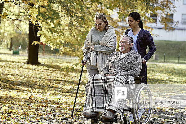 Gesundheitshelfer schiebt den Rollstuhl eines behinderten Mannes von einer älteren Frau  die mit Stock im Park geht