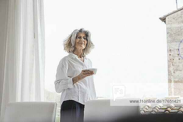 Smiling woman holding coffee cup standing by bright window at home