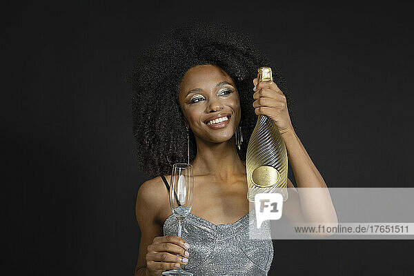 Smiling young woman holding champagne bottle and flute against black background