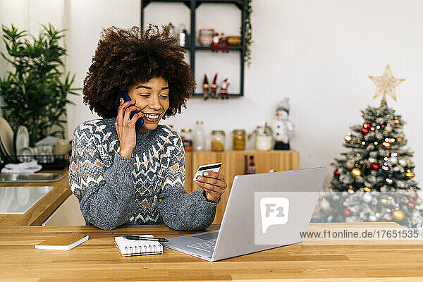Smiling young woman with credit card talking on mobile phone sitting at table