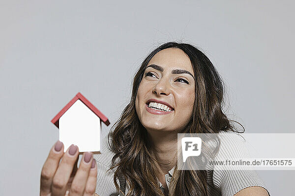 Fröhliche junge Frau hält ein Hausmodell vor grauem Hintergrund