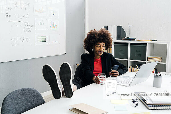Happy businesswoman sitting with feet up on desk and using mobile phone in office