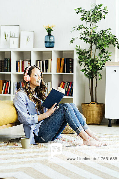 Smiling woman with headphones and book sitting by sofa on carpet at home