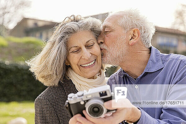 Senior man kissing woman taking selfie through camera on sunny day