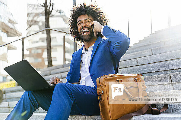 Happy businessman holding laptop talking on mobile phone sitting by shoulder bag