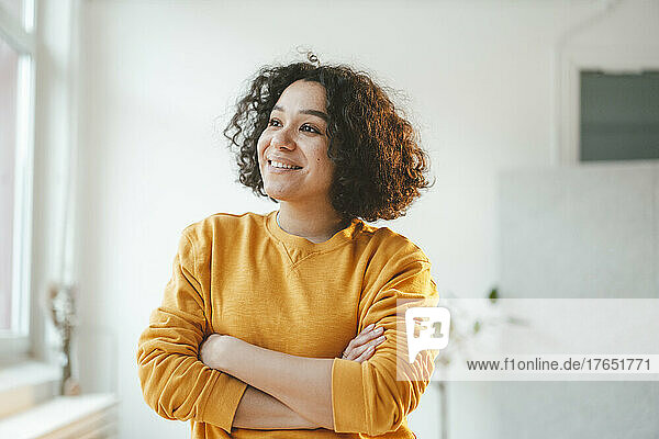 Smiling thoughtful woman standing with arms crossed at home