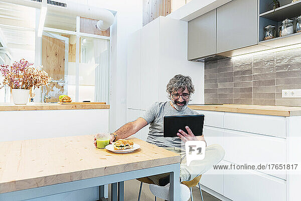 Mann sitzt mit Tablet-PC am Tisch und frühstückt zu Hause