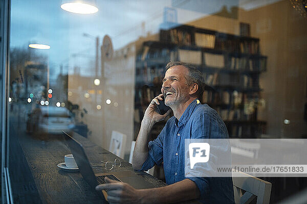 Happy freelancer talking on smart phone seen through glass window at cafe