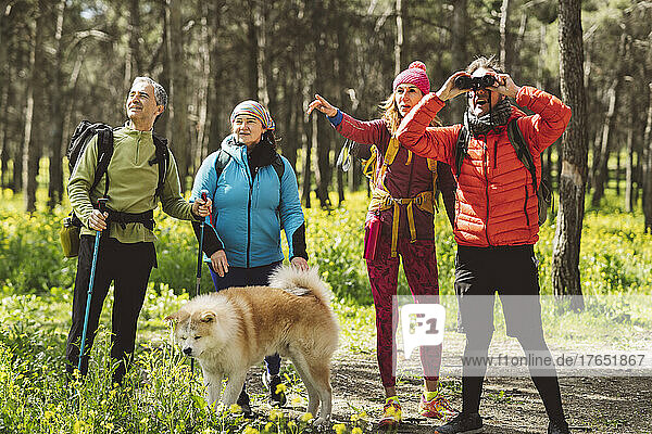 Mann schaut durch ein Fernglas und steht neben Freunden und Hund im Wald
