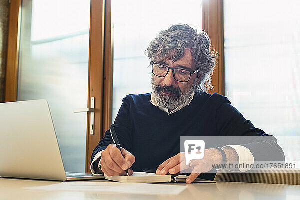 Freelancer writing in note pad sitting with laptop at table