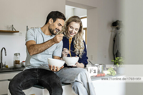 Happy couple eating noodles sitting on kitchen counter at home