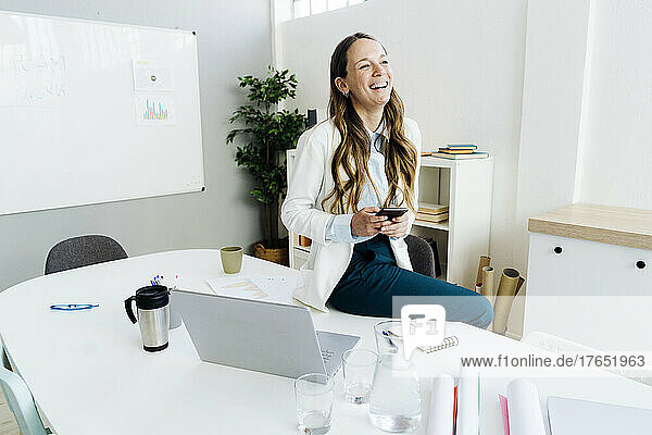 Happy businesswoman holding smart phone sitting on desk in office