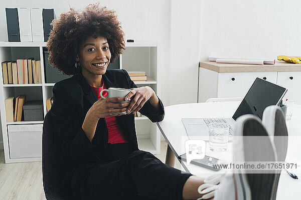 Smiling young businesswoman sitting at desk with coffee cup in office