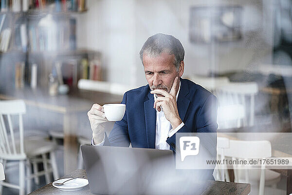 Businessman with hand on chin and coffee cup using laptop at cafe