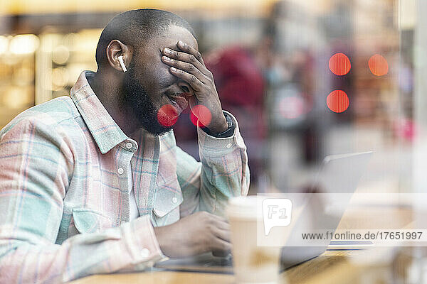 Worried freelancer with laptop sitting in cafe seen through glass