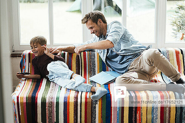 Happy father looking at playful son using smart phone sitting on sofa at home