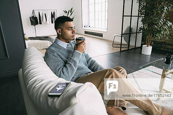 Man holding disposable coffee cup sitting by smart phone with flight mode symbol on sofa at home