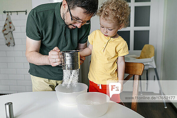 Man with daughter sieving flour at table in kitchen