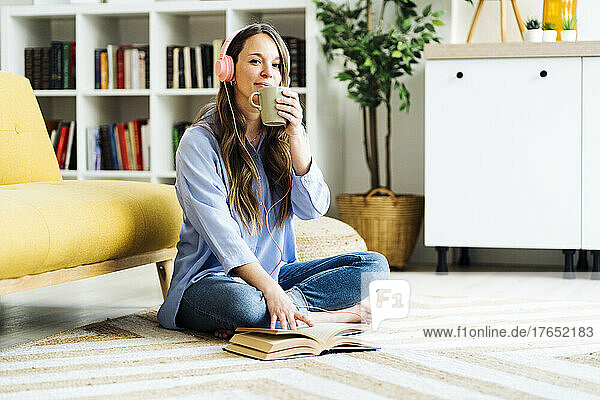 Woman having coffee sitting with book on carpet at home