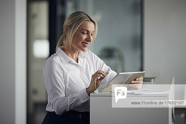 Smiling businesswoman using tablet PC standing by cabinet in office
