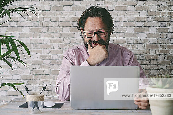 Lächelnder Geschäftsmann mit der Hand am Kinn und Blick auf den Laptop