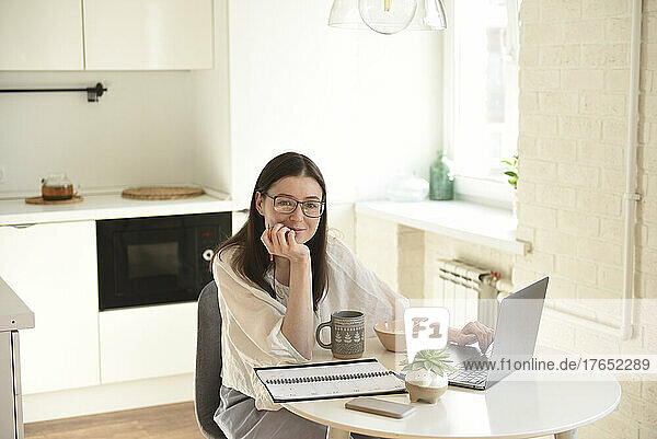 Smiling freelancer with laptop sitting at table in kitchen