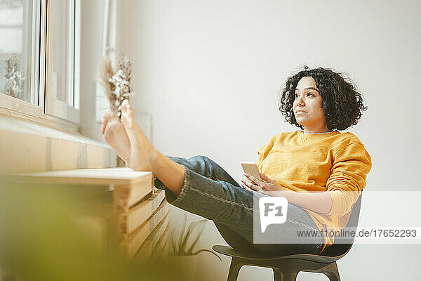 Thoughtful woman with smart phone sitting on chair at home