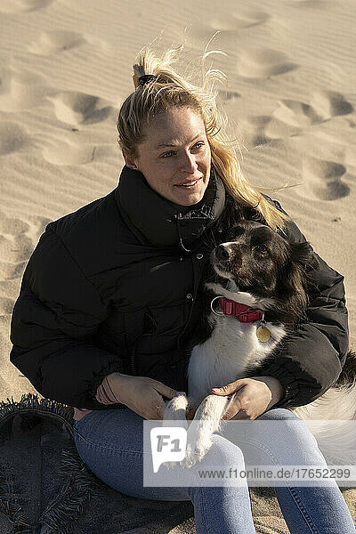 Smiling young blond woman sitting with pet dog on sand