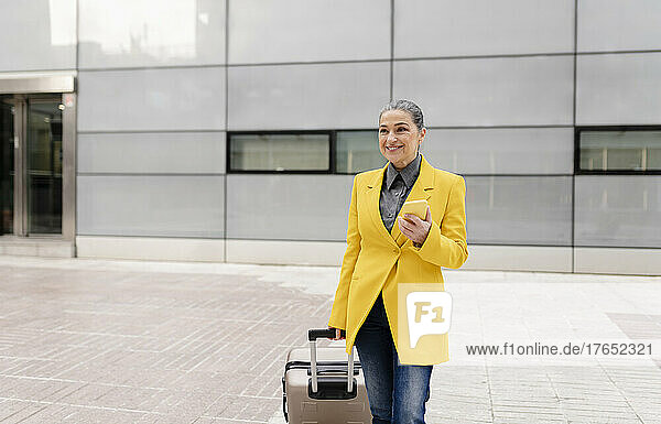 Smiling businesswoman holding smart phone walking with wheeled luggage on footpath