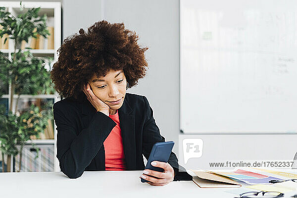 Bored businesswoman sitting at desk with hand on chin using mobile phone in office