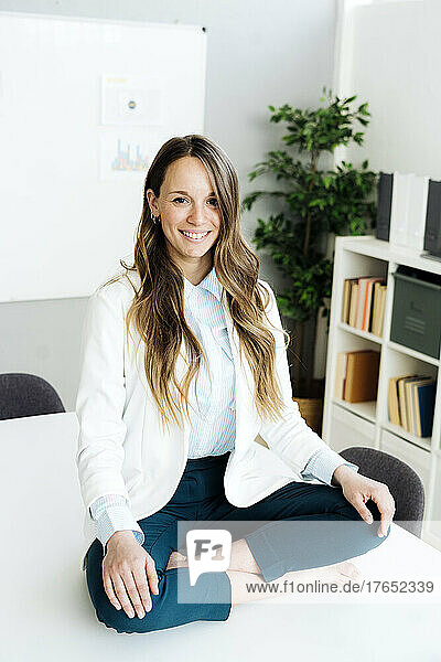 Happy businesswoman sitting cross-legged on desk in office